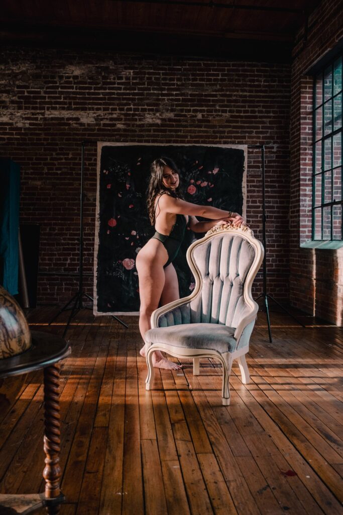Woman posing in front of painted canvas in green lingerie while leaning on a blue chair for a boudoir photography session