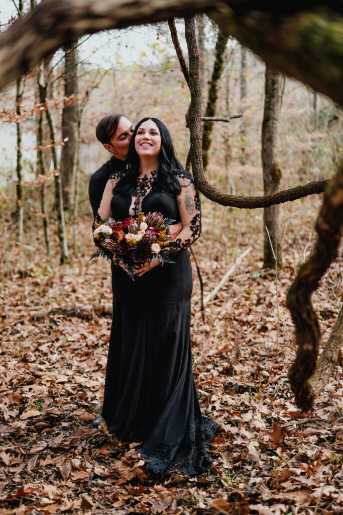Wedding couple wearing black embrace under vines in an open forest organized by wedding planner Rose and Ring Events